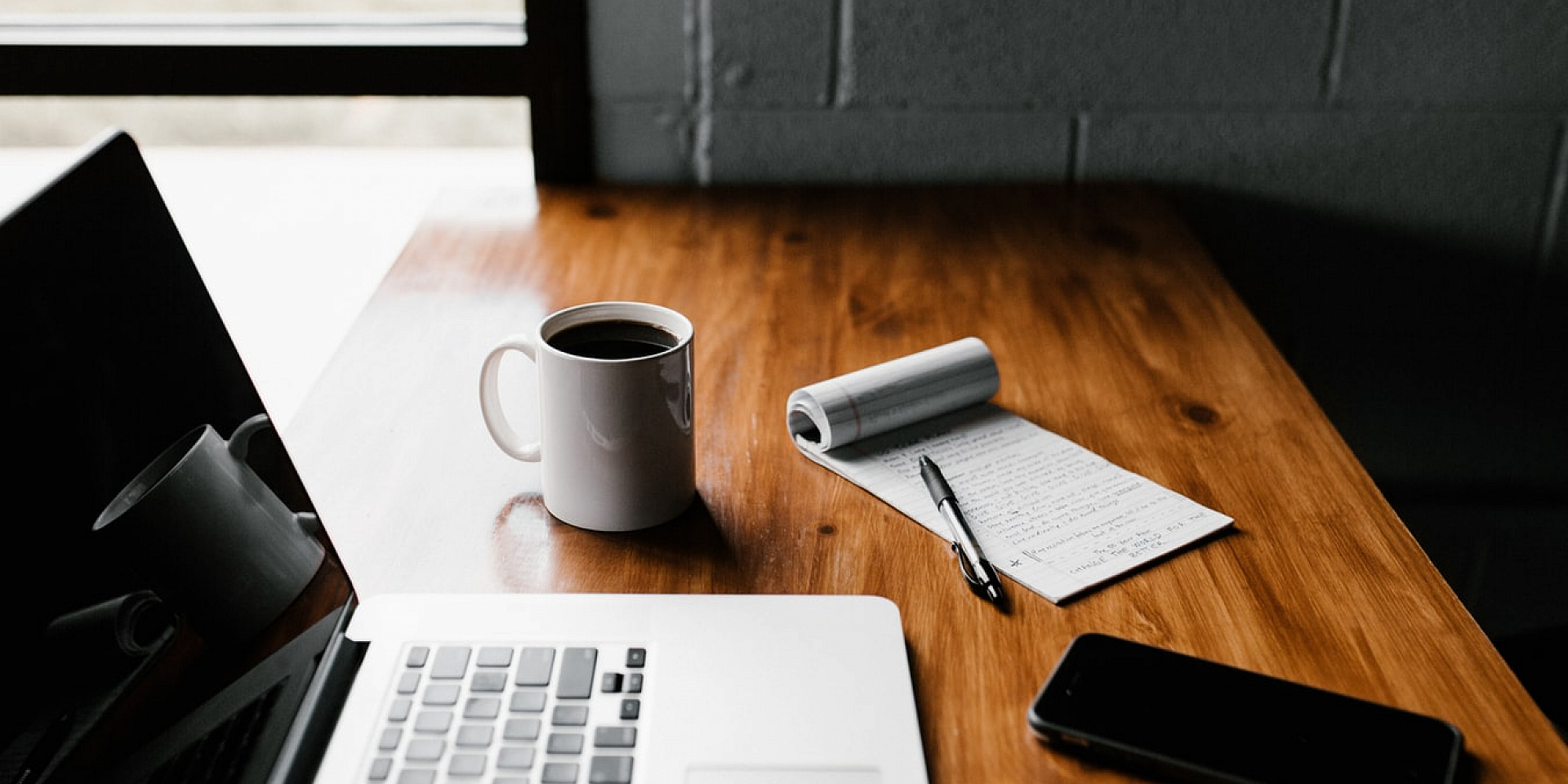 Laptop,coffee cup, notepad and cell phone on a table.