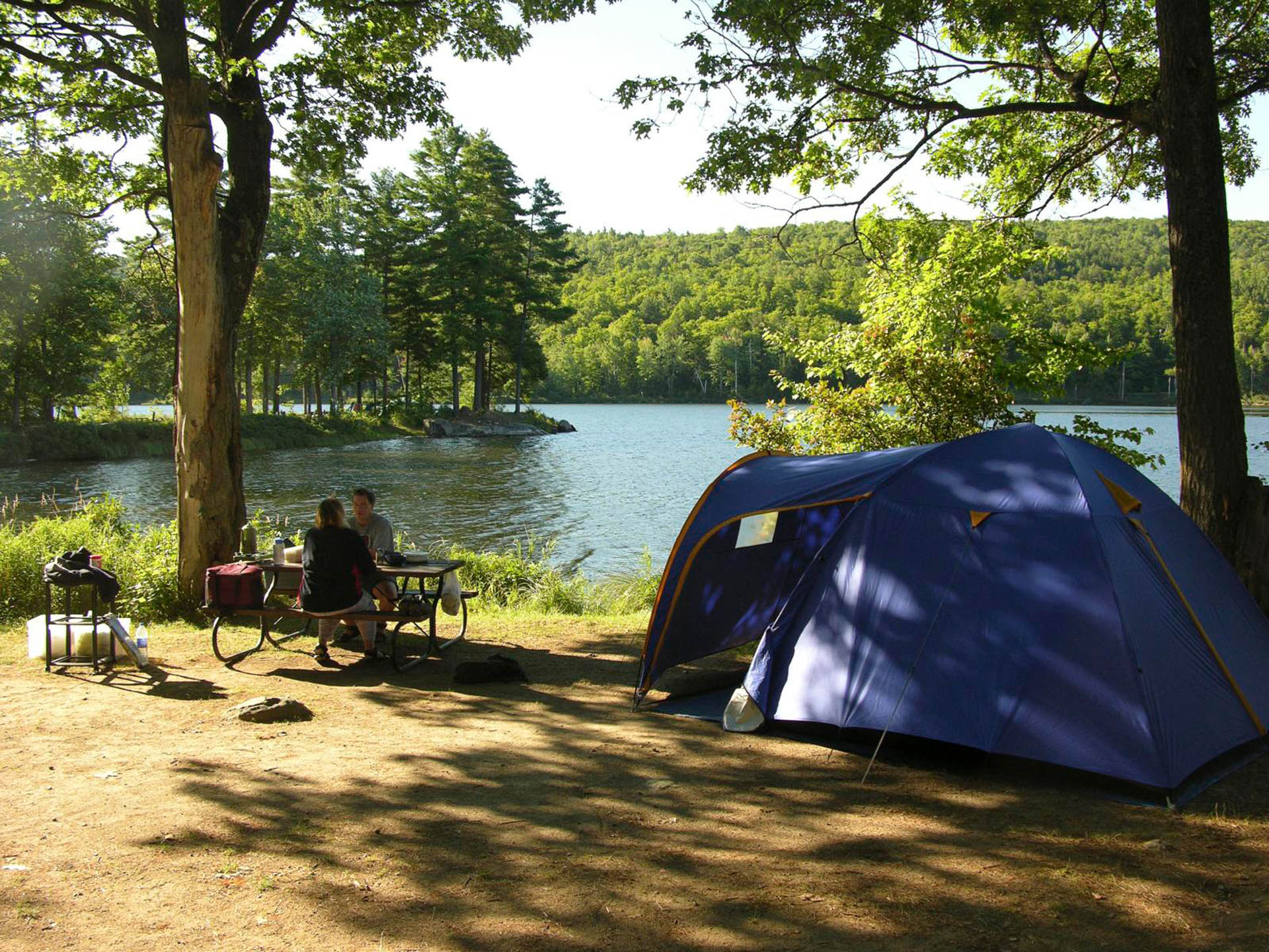 Couple sitting at a table near a tent on a camping site in the woods by a river