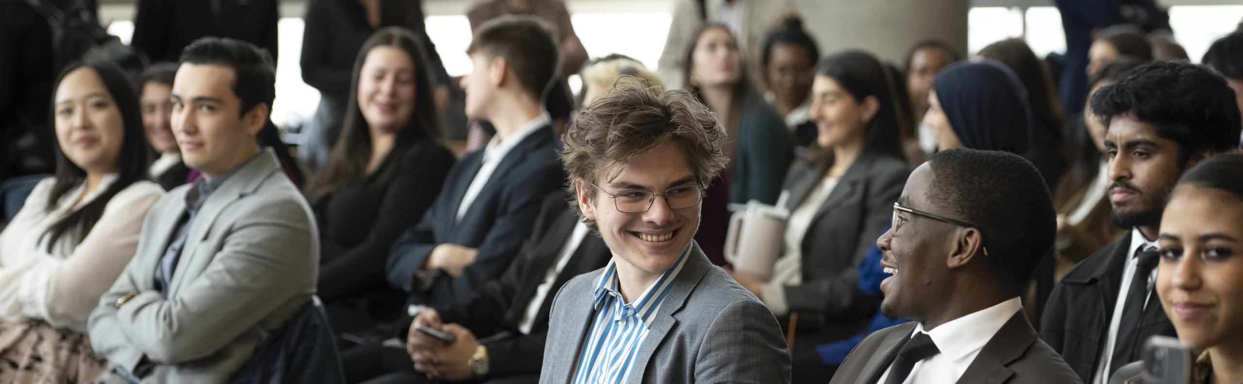 A diverse group of young professionals and students in an audience converse and smile at a formal event.