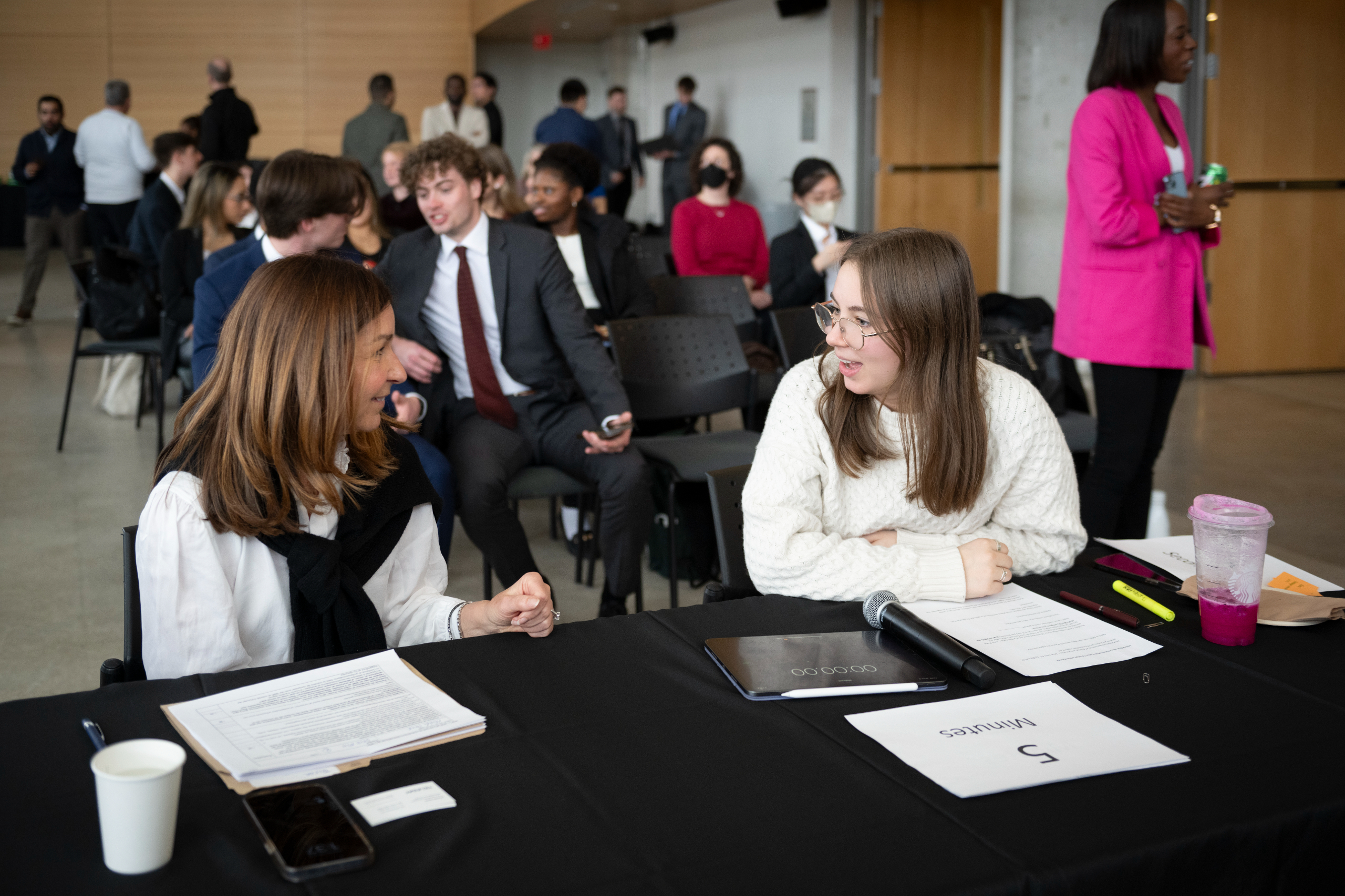 Two women sit at a judge's table at a case competition, conversing while participants sit behind them. 