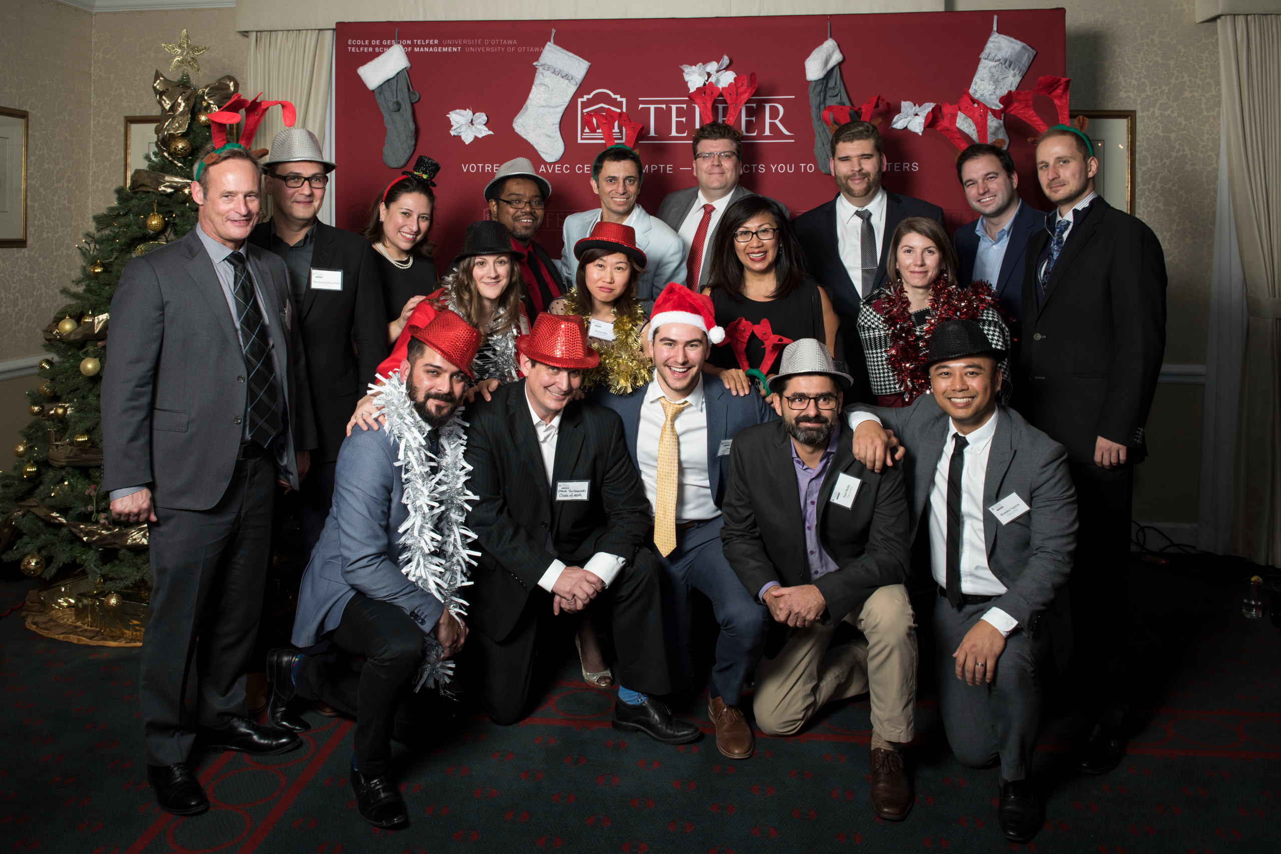 Original photo of a group Telfer EMBA alumni wearing holiday costume headwear posing in front of a burgundy Telfer backdrop at the annual holiday party