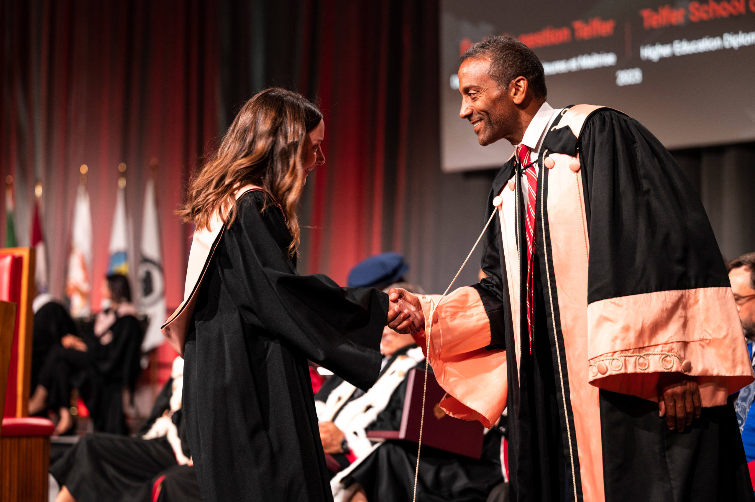 A graduate in a black gown shakes hands with Dean Stéphane Brutus, who is wearing a black and peach robe, on stage during convocation.