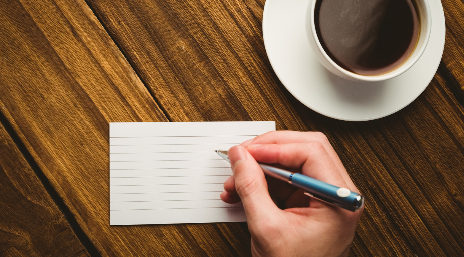 a hand hold a pen to start writing on the white flashcard on the desk, next to a cup of coffee