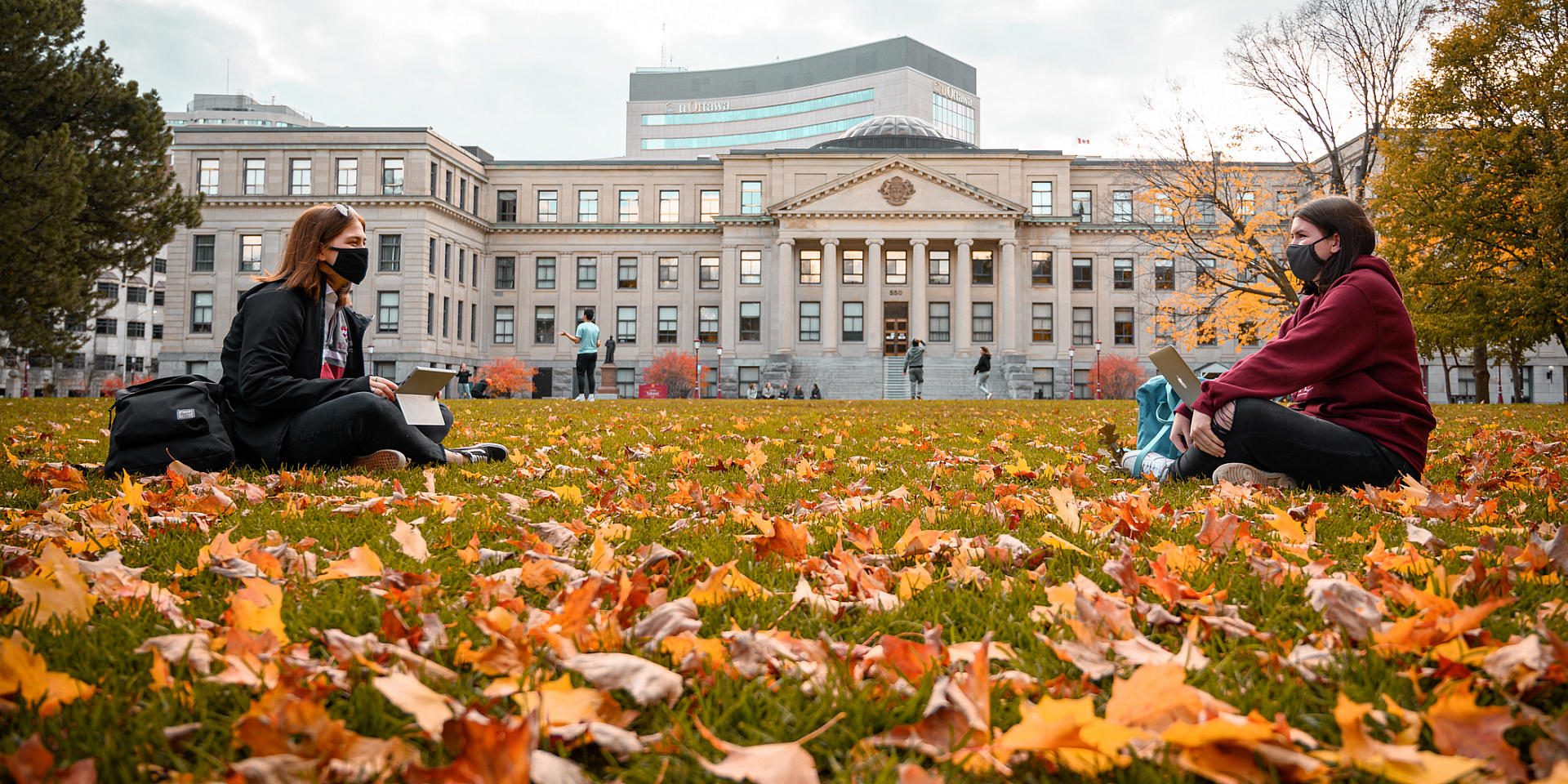 Two students study together in the field in front of the Tabaret building using their laptops