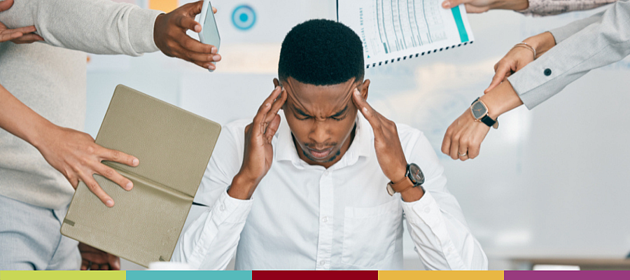 Worker at desk with his head in hands, while colleagues handing him several tasks to them