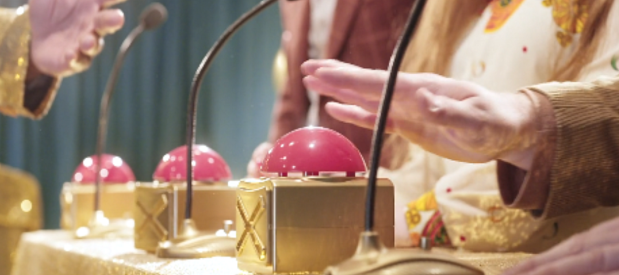 Participants pressing buzzers at a game-show-style table with microphones, representing a team-based quiz competition.