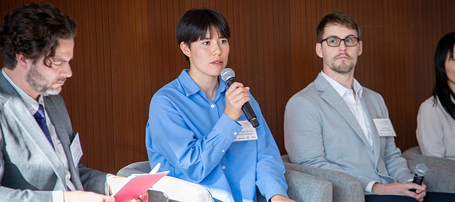 A panelist in a blue shirt speaks into a microphone while seated in an armchair during a sustainability discussion, alongside other panelists listening attentively.