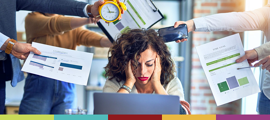 Worker at desk with head in hands, with colleagues handing several tasks to them